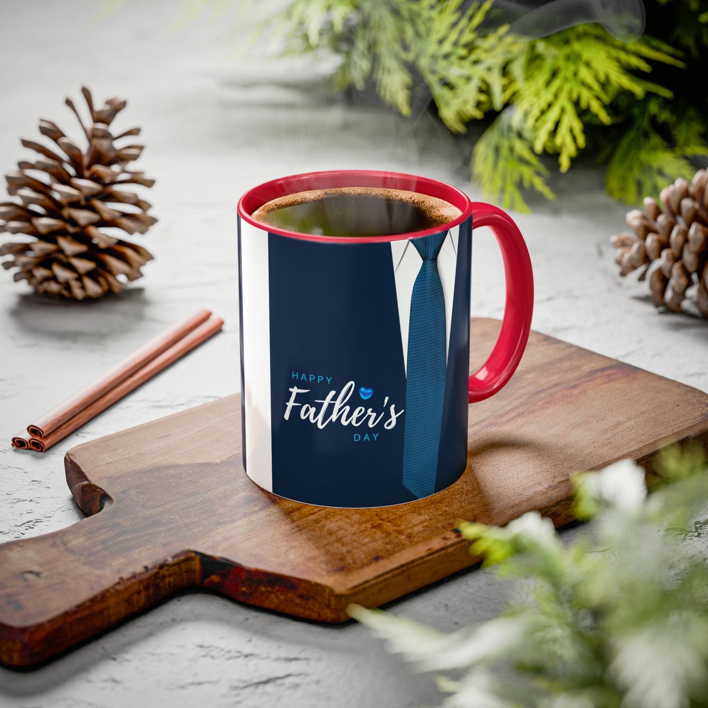 Colorful mugs with red interior and handle, featuring Father's Day design, on wooden board surrounded by greenery and pinecones.