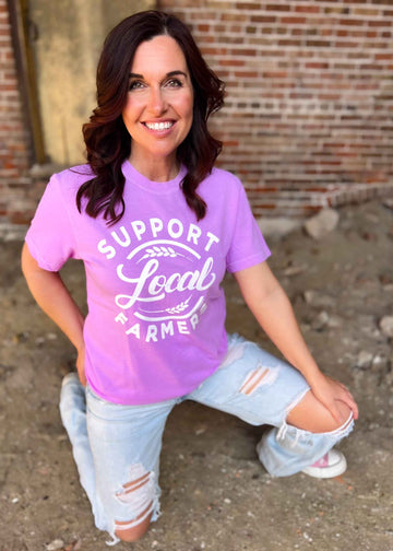 Neon violet graphic tee with "Support Local Farmers" design, worn by a smiling person kneeling, showcasing relaxed fit and crew neck style.