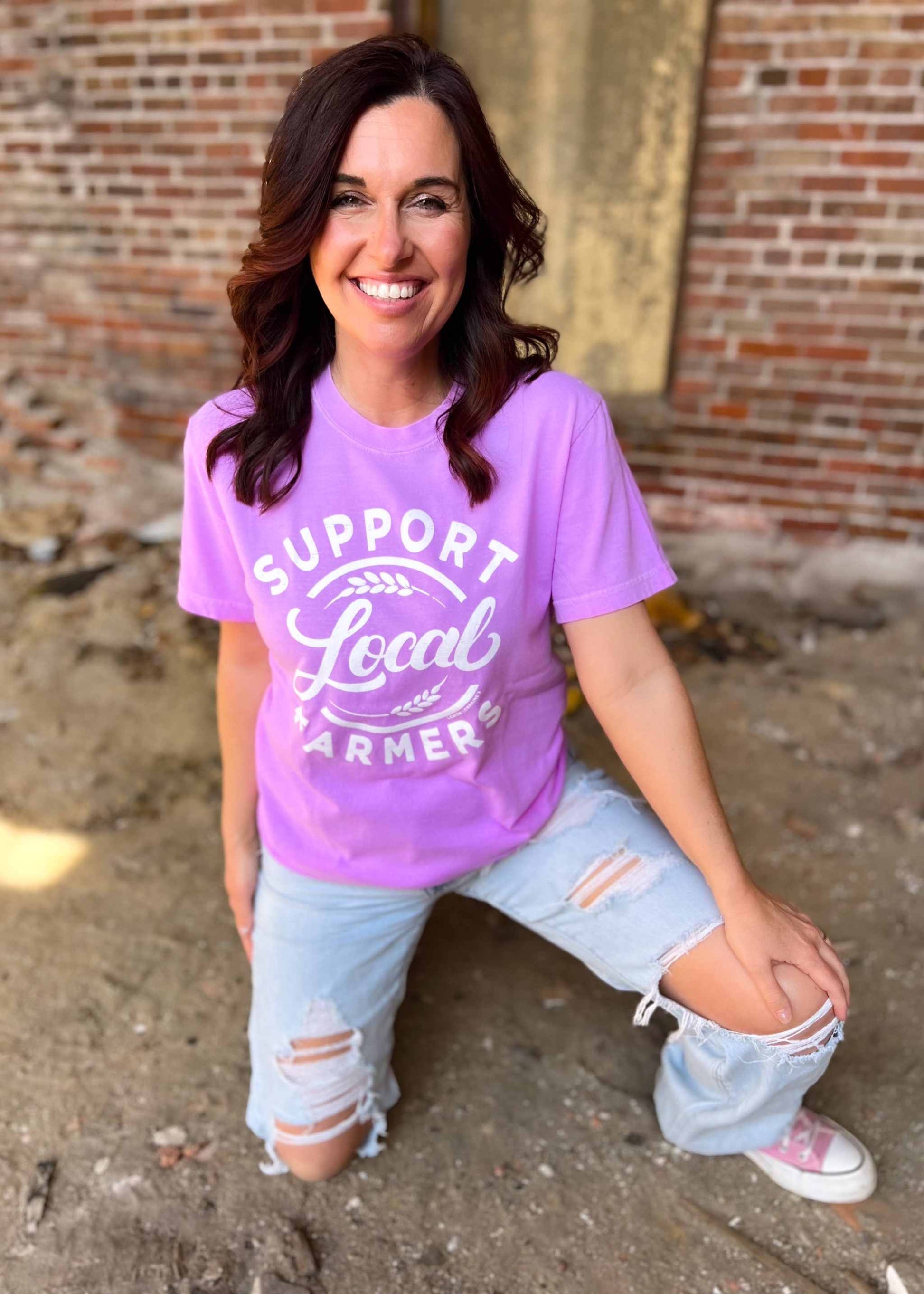 Neon violet graphic tee with "SUPPORT LOCAL FARMERS" print, worn by a person outdoors.