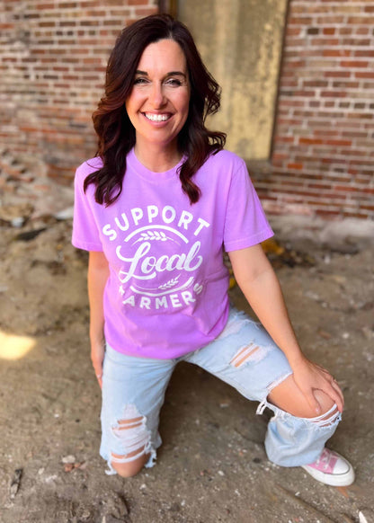 Neon violet graphic tee with "SUPPORT LOCAL FARMERS" print, worn by a person outdoors.