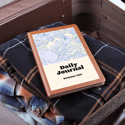 Daily Reflections journal with a floral design on a cozy plaid garment in a suitcase.