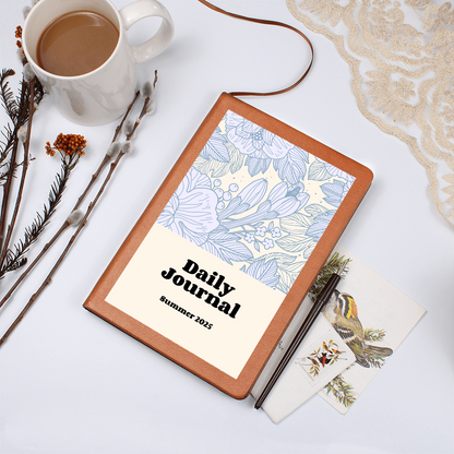 Daily Reflections journal with vegan leather cover on a table with coffee, pen, and postcard.